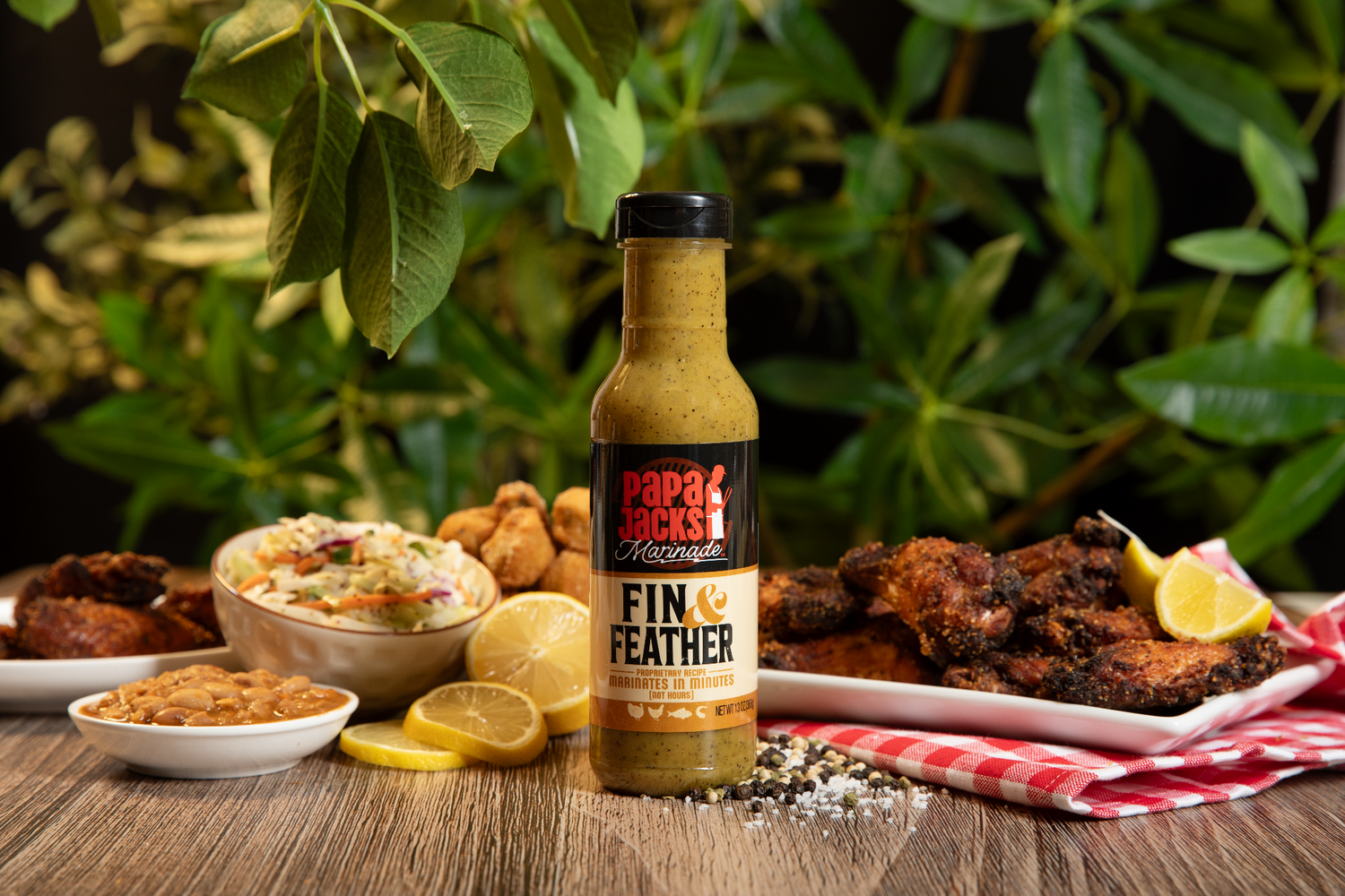 A bottle of Papa Jack’s Marinade Fin & Feather sauce stands in the center of a rustic wooden table, surrounded by a spread of crispy fried chicken, coleslaw, baked beans, hush puppies, and lemon slices. The background features lush green foliage, enhancing the fresh and flavorful appeal of the marinade.