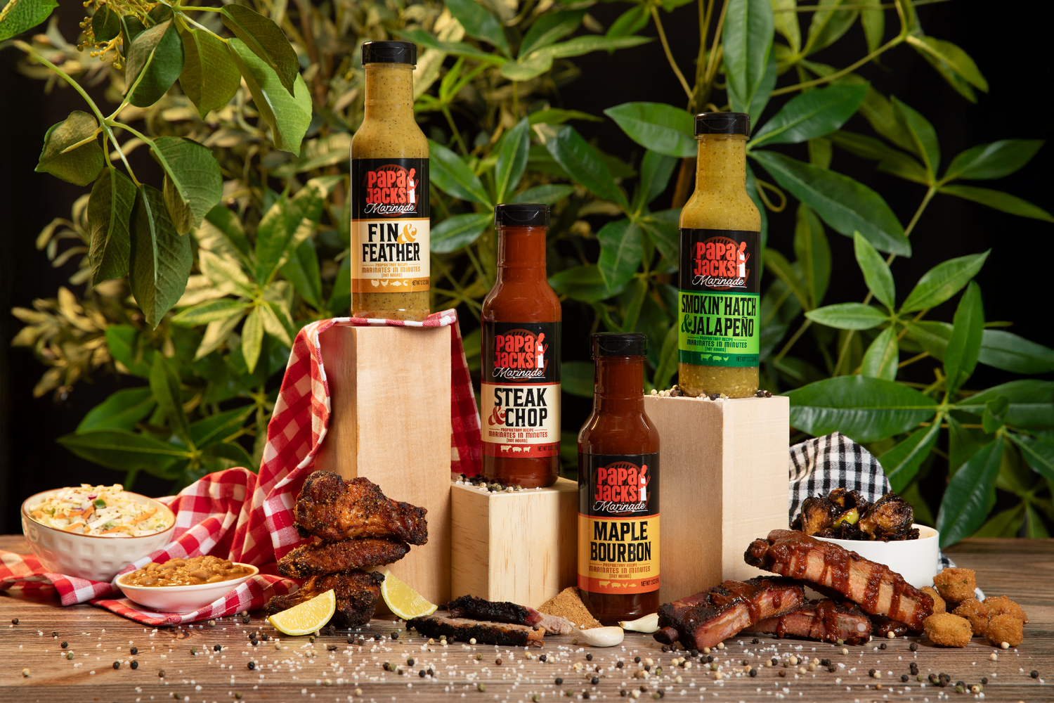 A full collection of Papa Jack’s Marinade sauces is elegantly displayed on a rustic wooden table, featuring four signature flavors: Fin & Feather, Steak & Chop, Maple Bourbon, and Smokin’ Hatch & Jalapeño. The bottles are arranged on wooden blocks with a red checkered cloth, surrounded by a mouthwatering spread of grilled ribs, chicken wings, coleslaw, baked beans, crispy hush puppies, and lemon wedges. The background is lush with greenery, adding a fresh and inviting feel to the scene.